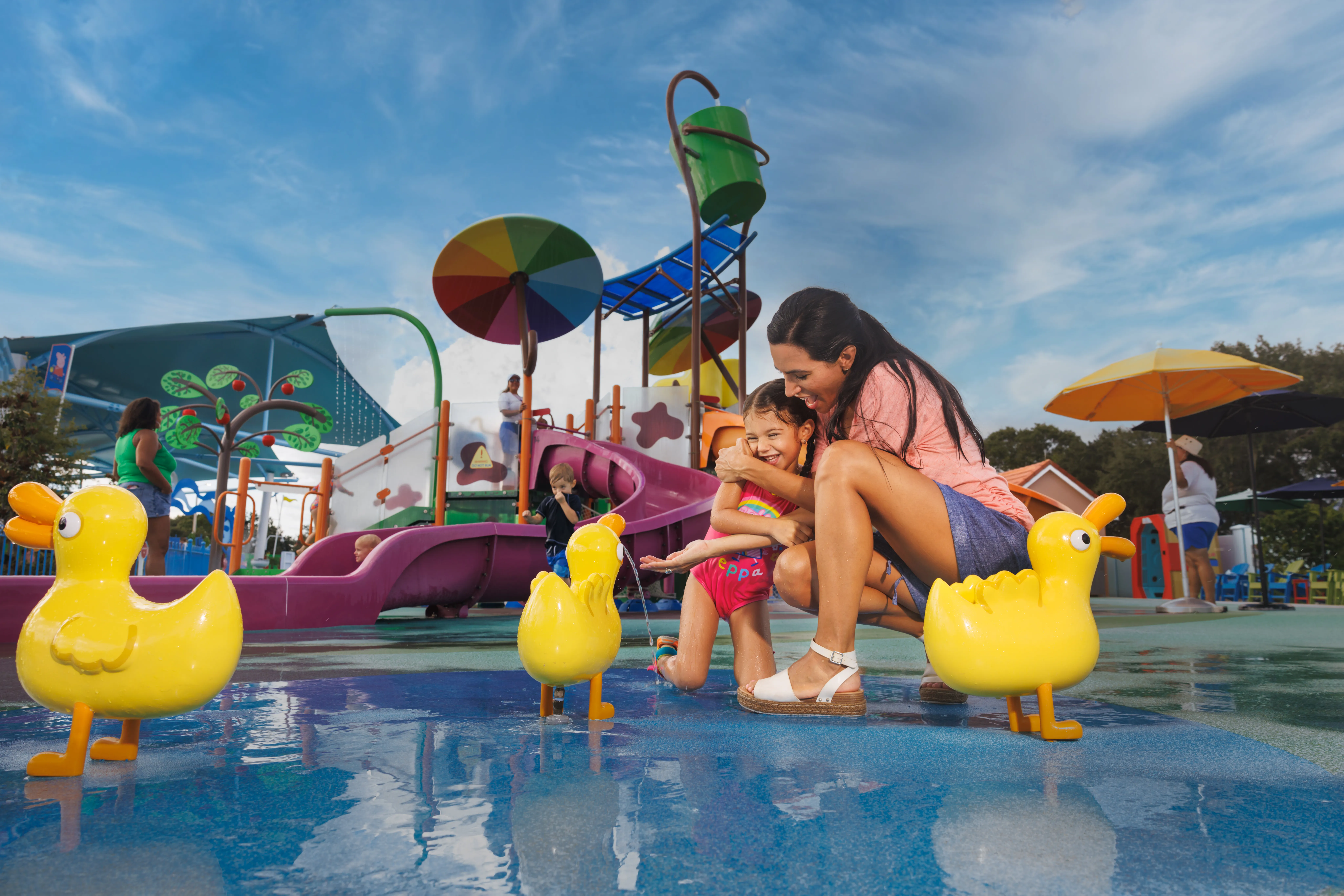 Una madre y su hija disfrutan de un momento divertido juntas en el Muddy Puddles Splash Pad en Peppa Pig Theme Park en Dallas Fort Worth. La hija salpica el agua mientras la madre observa, ambas sonriendo y riendo en esta atracción llena de agua y diversión.