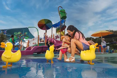 A mom and daughter enjoy a fun moment together at the Muddy Puddles Splash Pad at Peppa Pig Theme Park in Dallas Fort Worth. The daughter splashes in the water while the mom watches, both smiling and laughing in the playful, water-filled attraction.