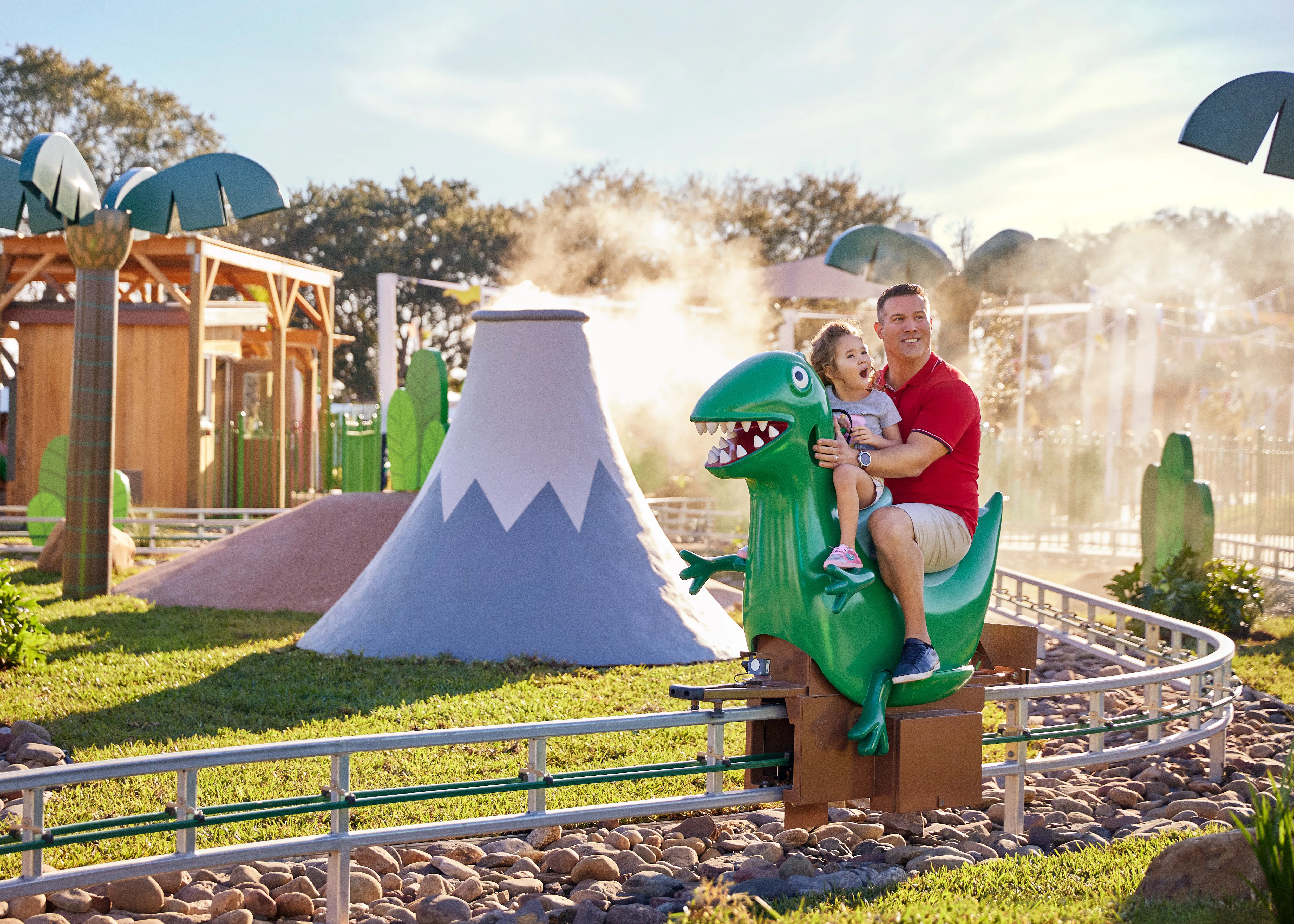 Un papá y su hija disfrutan montando la atracción Dinosaur Adventure de Grampy Rabbit en el Peppa Pig Theme Park en Dallas Fort Worth. Sonríen y se ríen mientras se embarcan en la emocionante atracción con temática prehistórica, rodeados por el colorido y divertido ambiente del parque.