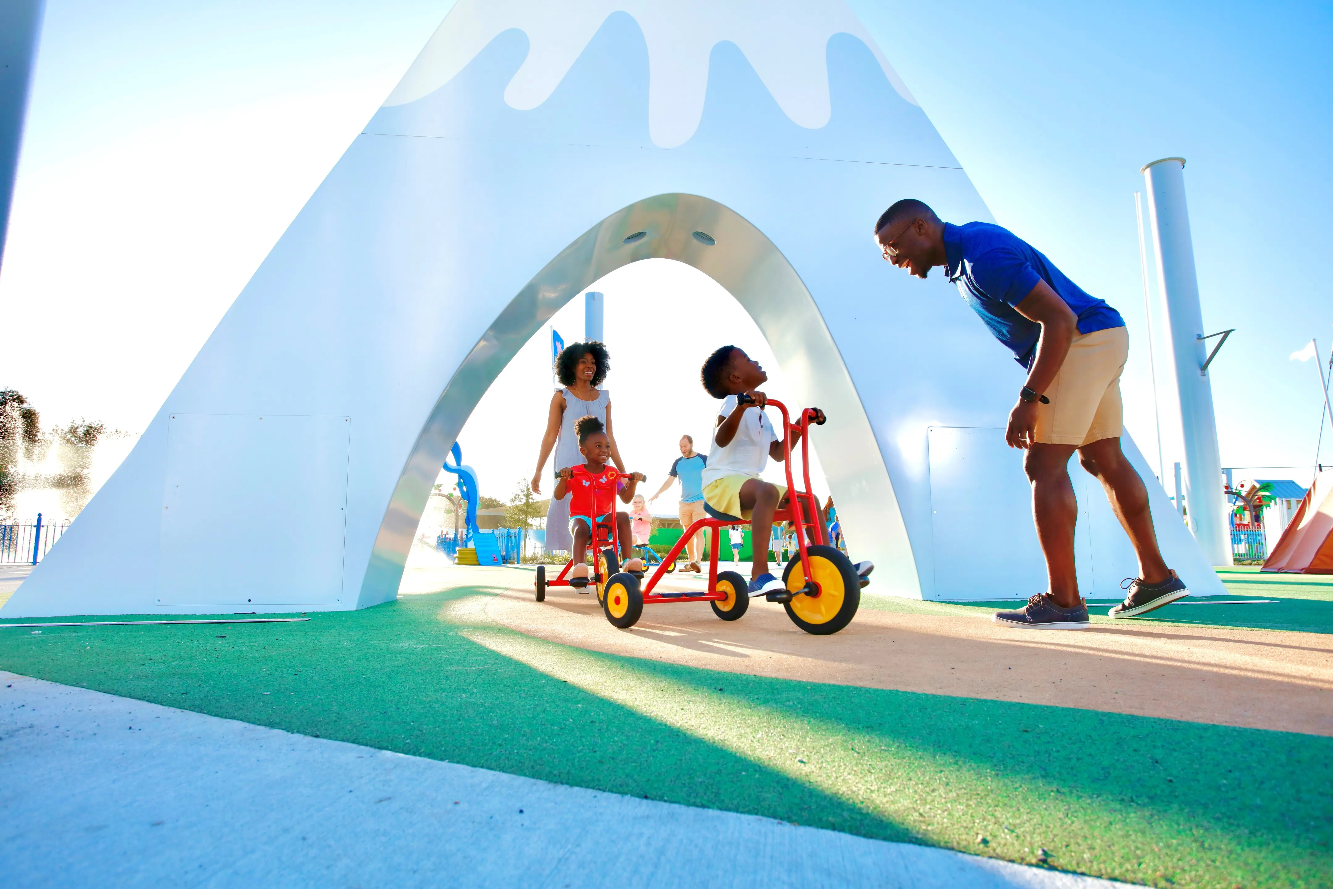 Una familia disfruta viendo a su hijos pedalear a través de Peppa’s Pedal Bike Tour y George’s Tricycle Trail en Peppa Pig Theme Park en Dallas Fort Worth. El niño pedalea felizmente a lo largo de los coloridos caminos, mientras los padres sonríen y animan, disfrutando de la divertida y pintoresca atmósfera del parque.
