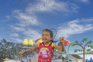 Una niña salpica agua en el Muddy Puddles Splash Pad en Peppa Pig Theme Park en Dallas Fort Worth. Ella sonríe y disfruta de las divertidas características acuáticas, rodeada de la colorida y alegre atmósfera del parque.