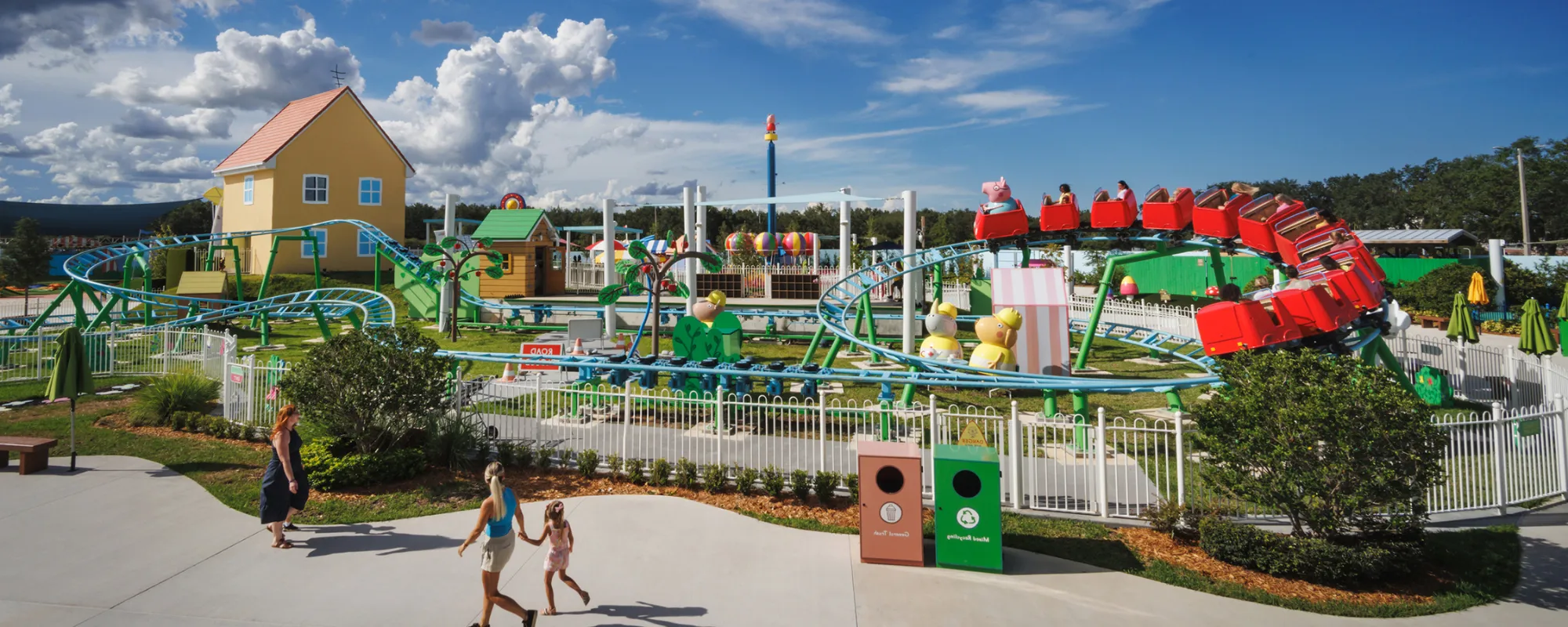 Los visitantes caminan por el Peppa Pig Theme Park en Dallas Fort Worth, disfrutando de la vibrante atmósfera. Al fondo, se puede ver la montaña rusa de Daddy Pig, añadiendo emoción a la experiencia llena de diversión.
