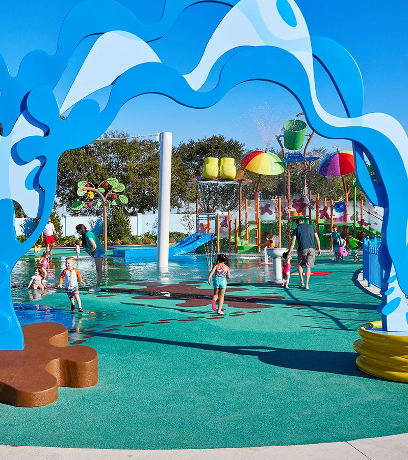 Niños y padres disfrutan del área de chapoteo Muddy Puddles en Peppa Pig Theme Park en Dallas Fort Worth. Los niños salpican el agua mientras los padres observan y se ríen, todo rodeado por la atmósfera juguetona y llena de agua del parque.