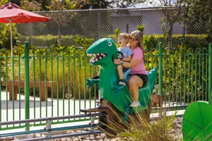 Una madre y su hijo disfrutan montando la atracción Dinosaur Adventure de Grampy Rabbit en el Peppa Pig Theme Park en Dallas Fort Worth. Sonríen y se ríen mientras se embarcan en la emocionante atracción con temática prehistórica, rodeados por el colorido y divertido ambiente del parque.