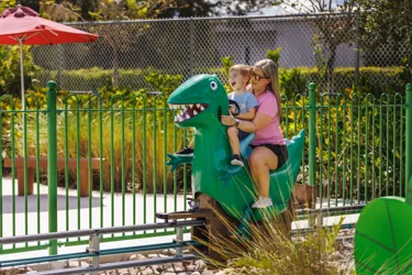 A mother and son enjoy riding Grampy Rabbit's Dinosaur Adventure at Peppa Pig Theme Park in Dallas Fort Worth. They smile and laugh as they embark on the exciting, prehistoric-themed ride, surrounded by the colorful and fun atmosphere of the park.