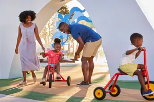 Una familia disfruta viendo a su hijos pedalear a través de Peppa’s Pedal Bike Tour y George’s Tricycle Trail en Peppa Pig Theme Park en Dallas Fort Worth. El niño pedalea felizmente a lo largo de los coloridos caminos, mientras los padres sonríen y animan, disfrutando de la divertida y pintoresca atmósfera del parque.